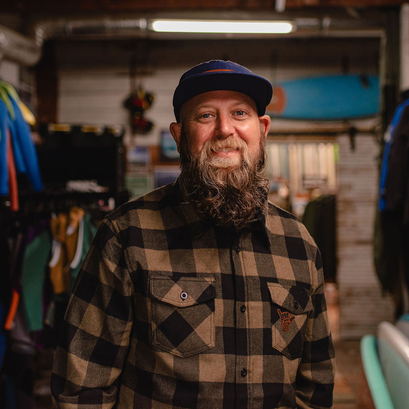 Load image into Gallery viewer, A smiling man with a full beard wearing the Shaka Flannel and a dark blue cap, standing in a shop interior with surfboards and clothing racks in the background.
