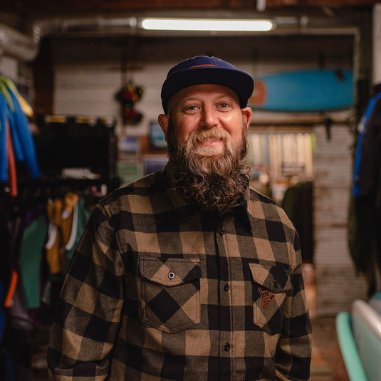 A smiling man with a full beard wearing the Shaka Flannel and a dark blue cap, standing in a shop interior with surfboards and clothing racks in the background.