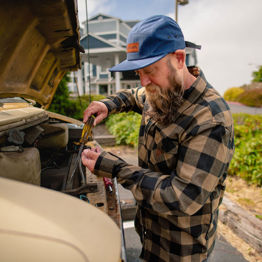 A bearded man wearing a blue cap and the Shaka Flannel leans over an open car hood, working on the engine outdoors.