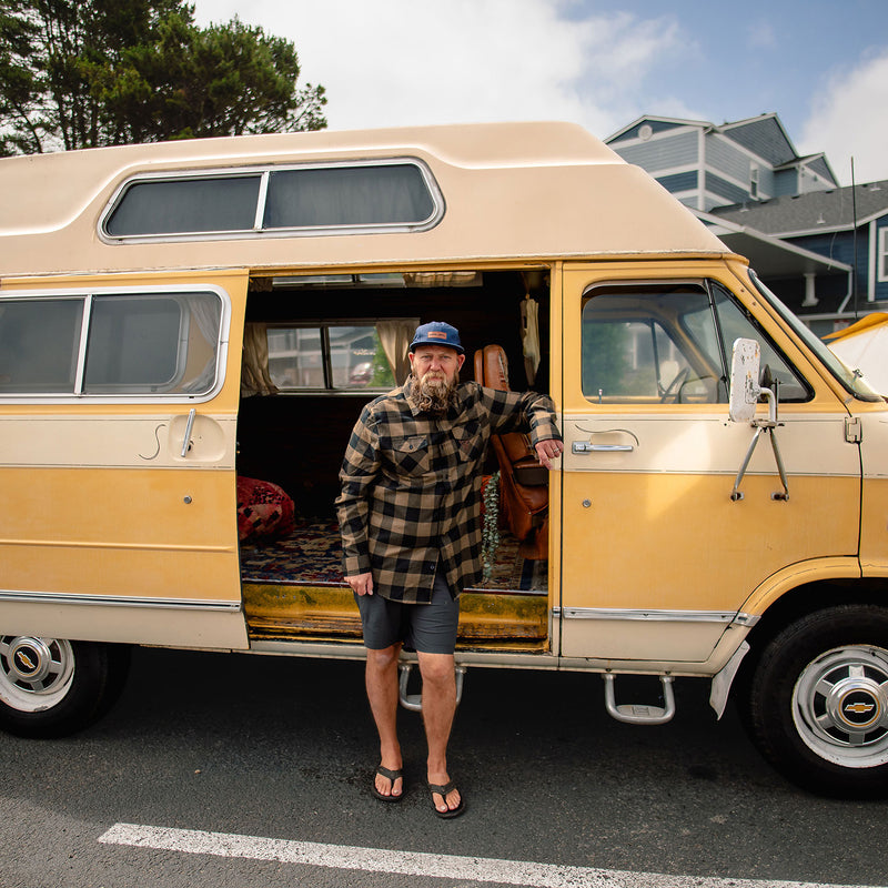 Load image into Gallery viewer, A man wearing a blue cap, the Shaka Flannel, and dark shorts stands in front of an open side door of a vintage two-tone beige and yellow camper van parked outdoors on a street.

