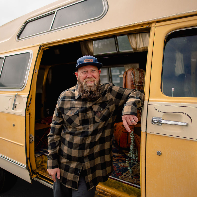 Load image into Gallery viewer, A bearded man wearing a blue cap and the Shaka Flannel leans against the open door of a vintage-style yellow camper van.
