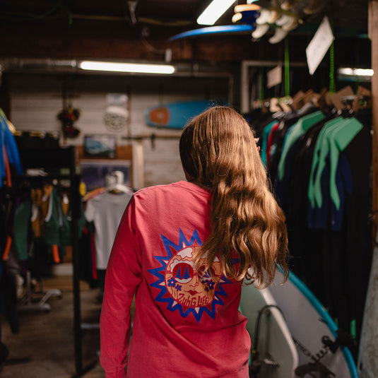 A person standing in a retail store, viewed from behind, wearing the Sun Kissed Long Sleeve T-Shirt with a large blue and red graphic print on the back.