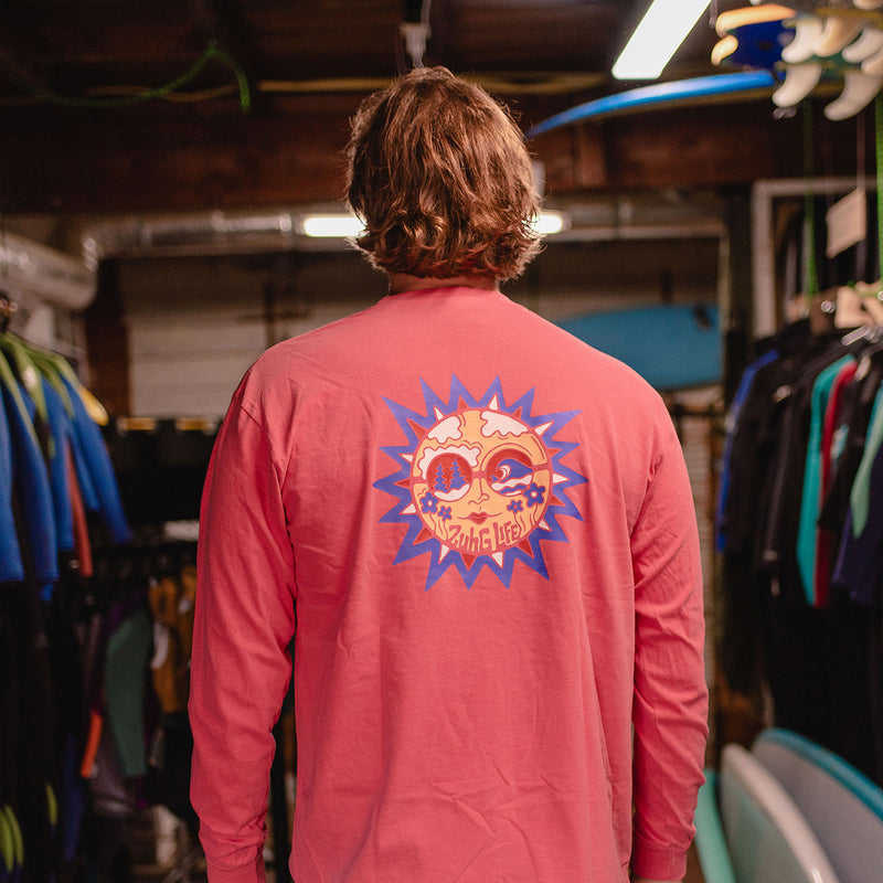 Load image into Gallery viewer, A person viewed from behind wearing the Sun Kissed Long Sleeve T-Shirt with a large colorful sun graphic on the back, standing in a retail or workshop environment with surfboards and racks visible.
