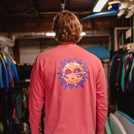 A person viewed from behind wearing the Sun Kissed Long Sleeve T-Shirt with a large colorful sun graphic on the back, standing in a retail or workshop environment with surfboards and racks visible.
