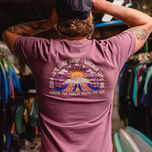 A man viewed from behind wearing the Where The Forest Meets The Sea T-Shirt with a large graphic print on the back. The graphic features a sunset, waves, a sailboat, and the text 'ZUIG LIFE SURF SHOP - LINCOLN CITY OR WHERE THE FOREST MEETS THE SEA'.