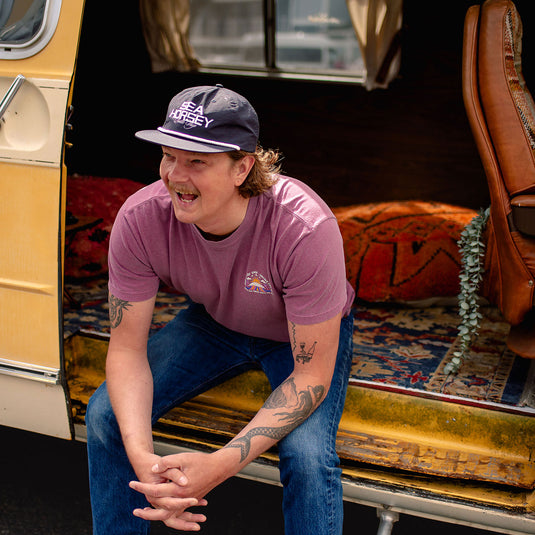 A man wearing the Where The Forest Meets The Sea T-Shirt, blue jeans, and a dark navy baseball cap with 'PEAK HARLEY' printed on the front, sitting inside the open back of a vintage cream-colored van with patterned rugs and leather seats.