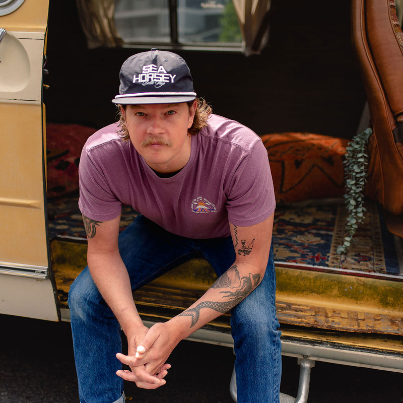 Load image into Gallery viewer, A man wearing the Where The Forest Meets The Sea T-Shirt, blue jeans, and a dark gray baseball cap.
